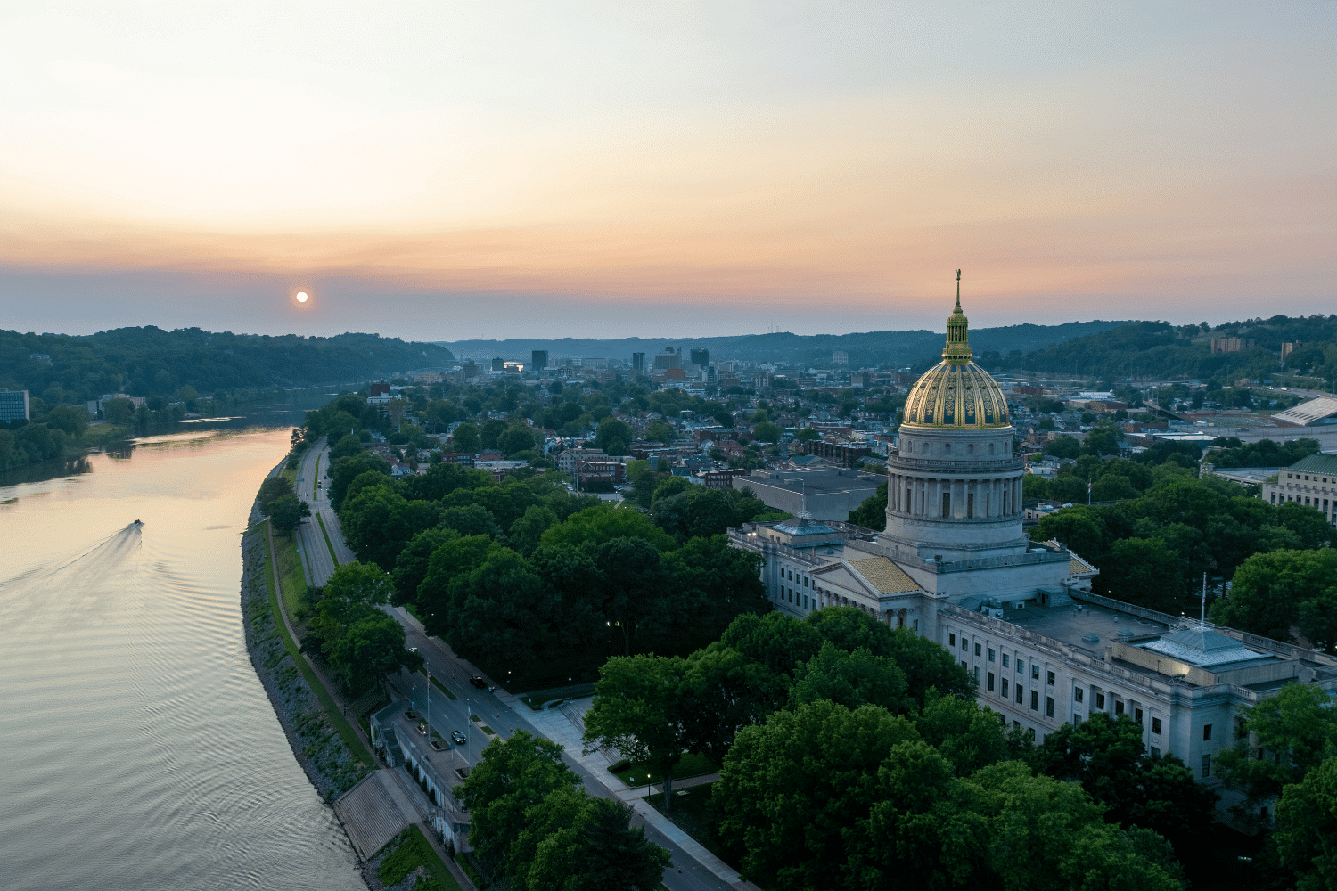 Aerial view of a domed government building, where debates over the West Virginia Machine Gun Bill unfold, surrounded by trees and a nearby river reflecting the setting sun as a boat leaves a wake under a hazy sky.