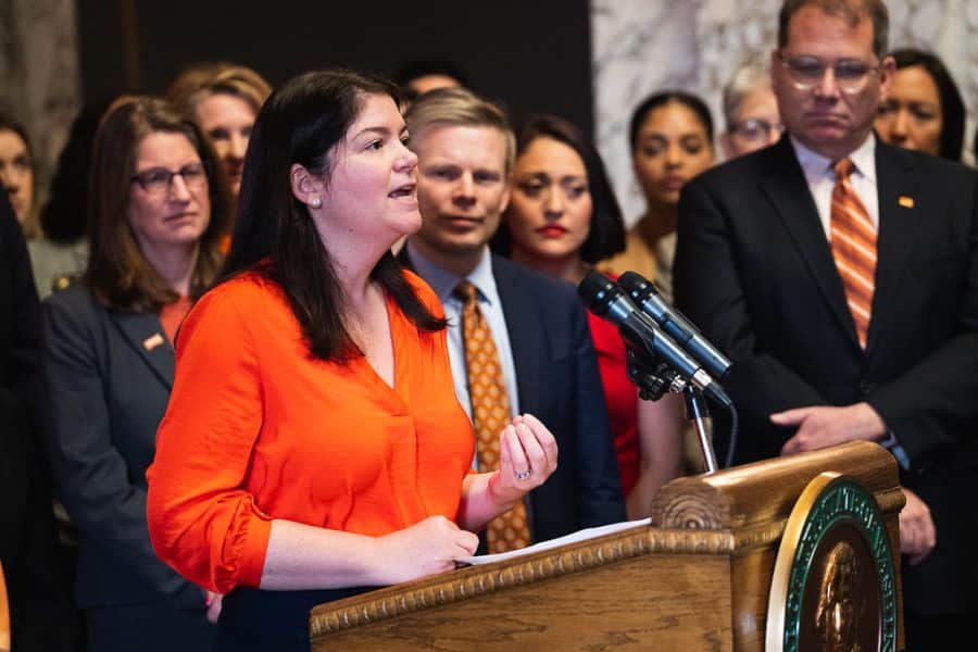 2A Marketplace: Private Firearm Classifieds A woman in an orange blouse speaks at a podium with microphones, surrounded by a group of people in formal attire, some wearing orange, in a government or official setting. Private sale of guns.
