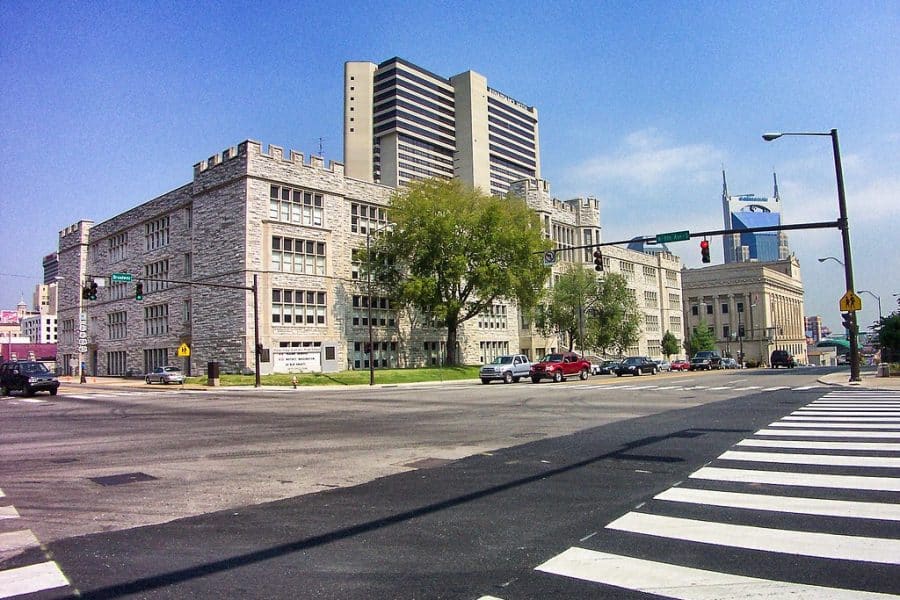 2A Marketplace: Private Firearm Classifieds A stone building with castle-like features sits on a city corner with cars parked nearby. Modern high-rises and a blue sky are visible in the background. A zebra-striped crosswalk and traffic lights are in the foreground. Private sale of guns.