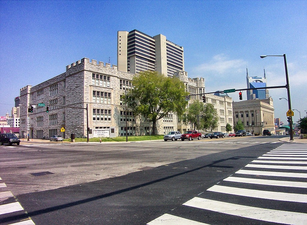2A Marketplace: Private Firearm Classifieds A stone building with castle-like features sits on a city corner with cars parked nearby. Modern high-rises and a blue sky are visible in the background. A zebra-striped crosswalk and traffic lights are in the foreground. Private sale of guns.