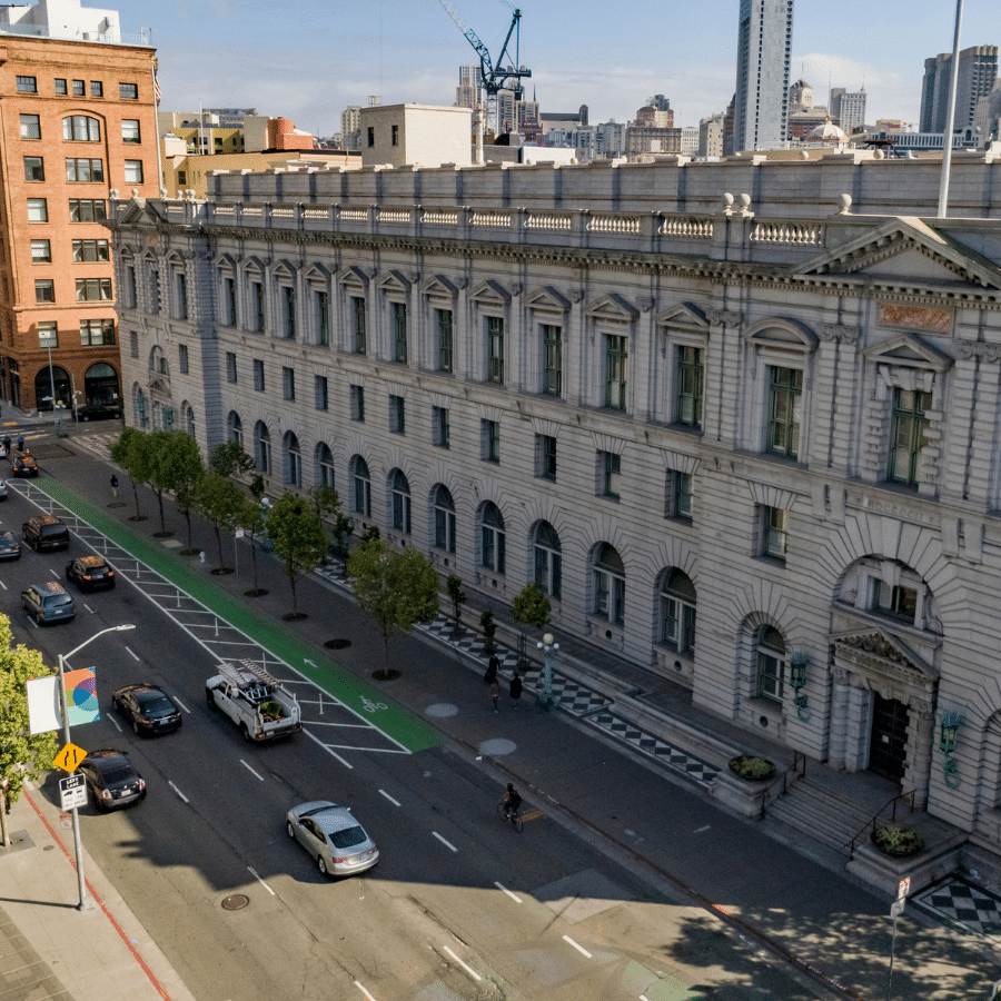 2A Marketplace: Private Firearm Classifieds A wide street runs beside a large, ornate stone building with arched windows and columns. Cars and a bike are on the road, and trees line the sidewalk. City buildings and a crane appear in the background, evoking an urban California landscape often discussed in recent news about large-capacity magazine confiscation laws. Private sale of guns.