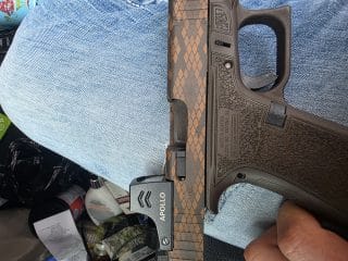 2A Marketplace: Private Firearm Classifieds A close-up of a custom-patterned Glock handgun with an APOLLO optic sight, held by someone wearing blue jeans. Various items, including papers and containers, are visible in the background. Private sale of guns.