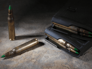 2A Marketplace: Private Firearm Classifieds Three rifle cartridges with green-tipped bullets are shown, with two magazines partially filled with similar rounds on a textured stone surface under strong light. Private sale of guns.