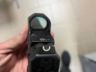 2A Marketplace: Private Firearm Classifieds Close-up view of a persons hand holding a black handgun with a visible reflex sight, aimed towards the floor with tiled flooring in the background. Private sale of guns.