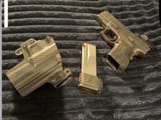 2A Marketplace: Private Firearm Classifieds A black handgun, a matching magazine, and a carbon-fiber-patterned holster are placed on a textured dark fabric surface. Private sale of guns.